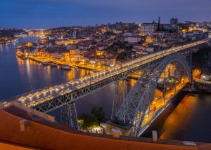 View of Porto’s iconic bridge spanning the Douro River, with colorful lights at dusk and a glimpse of the city’s hidden gems beyond the main tourist spots.